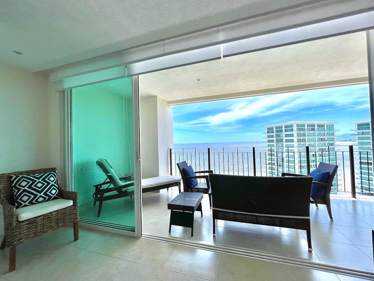 A bright seating area is visible, featuring a stylish wicker sofa and chairs with decorative cushions. Large glass doors open onto a spacious balcony with stunning water views. A lounge chair adds comfort, while a small coffee table sits in front, enhancing the space's functionality.