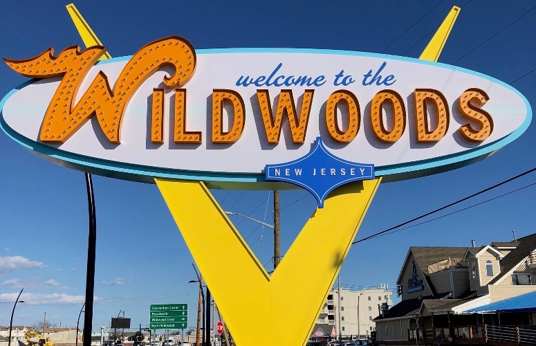 The iconic Welcome to the Wildwoods sign is prominently displayed against a clear blue sky. The sign features bold letters in orange and yellow hues, with a retro design that highlights the area's vibrant character. Nearby structures are visible, complementing the coastal atmosphere.