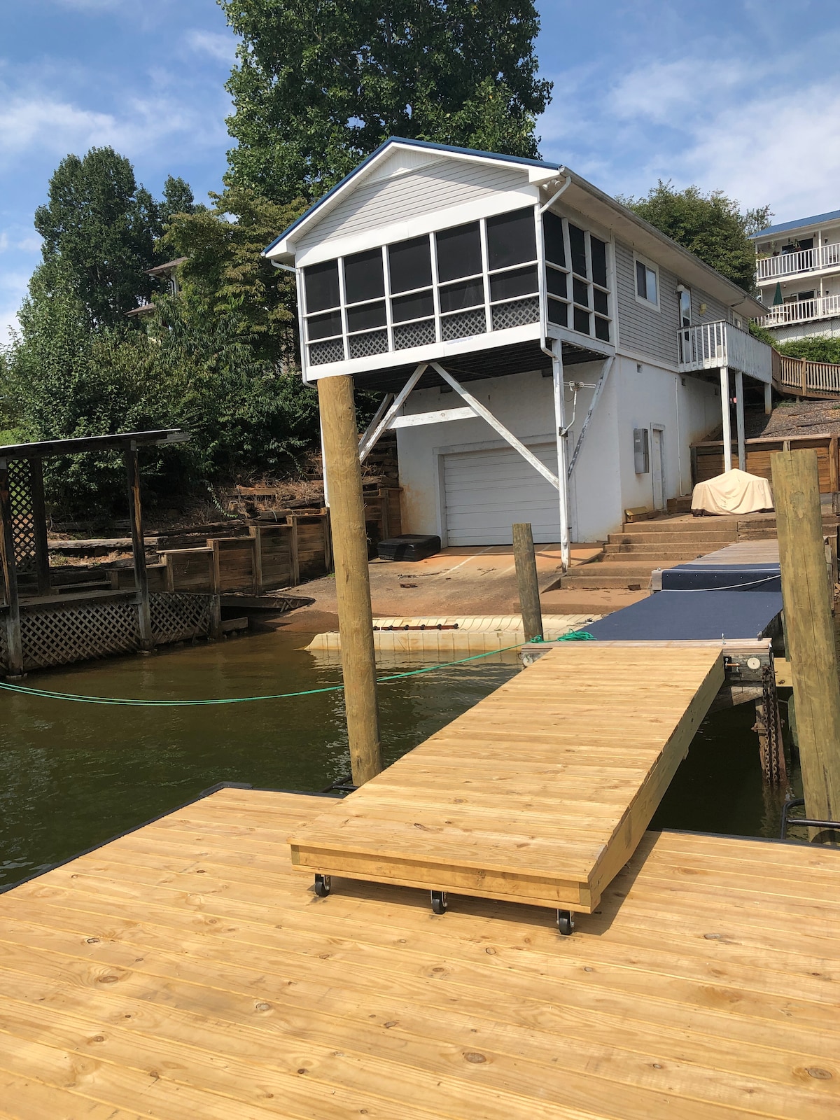 A wooden dock extends towards a charming boathouse positioned above the water. The boathouse is elevated with a screened-in porch, featuring a white exterior. Surrounding greenery adds a natural touch, while the calm water reflects a clear blue sky.