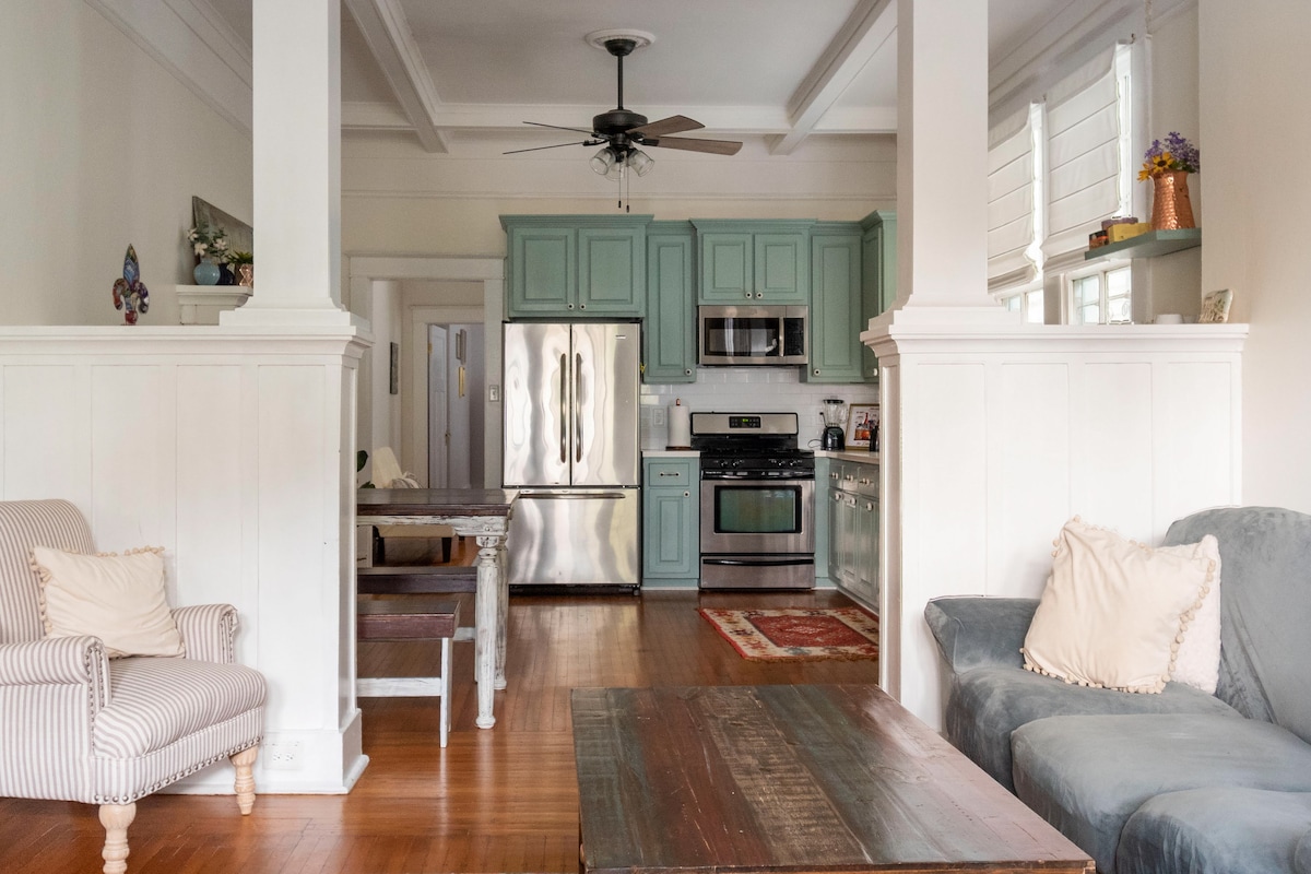 An open living area features a combination of light walls and wooden floors, complemented by two comfortable sofas. A well-equipped kitchen with stainless steel appliances and green cabinetry is visible, along with a dining table in the background, enhancing the spacious atmosphere.