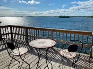 A private balcony features a round table with decorative patterns and two black iron chairs, overlooking tranquil water. The scene is complemented by a bright sky and distant greenery, providing a peaceful setting for relaxation.