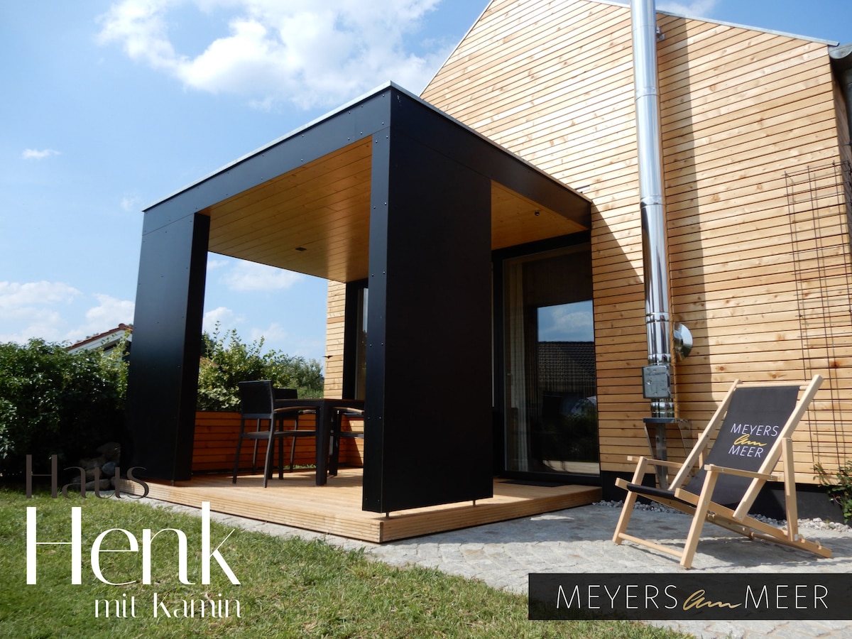 An outdoor terrace is featured, designed with a sleek black roof and wooden flooring. A dining table and chairs provide space for meals, while a deck chair adds a relaxed spot to enjoy the surroundings. The exterior is clad in natural wood, enhancing a harmonious atmosphere.