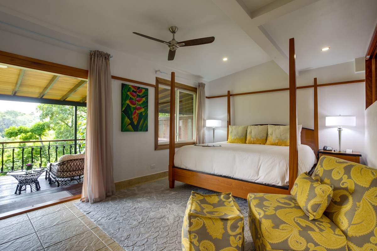 A cozy bedroom features a four-poster bed with yellow accent pillows and a patterned area rug. Natural light fills the space from large windows, revealing a balcony with outdoor seating overlooking lush greenery.