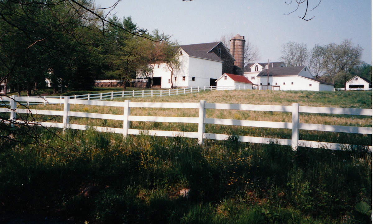 A sprawling landscape showcases a historic farmstead with white buildings and a tall silo along a grassy field. A white fence lines the property, surrounded by trees and open fields, conveying a serene rural atmosphere.