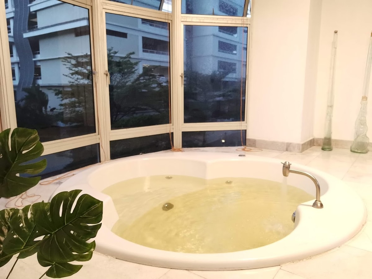 A round jacuzzi is positioned beside large windows, allowing natural light to enter the space. The water appears slightly tinted, and subtle textures can be seen in the marble flooring. A green plant adds a touch of greenery near the jacuzzi's edge.