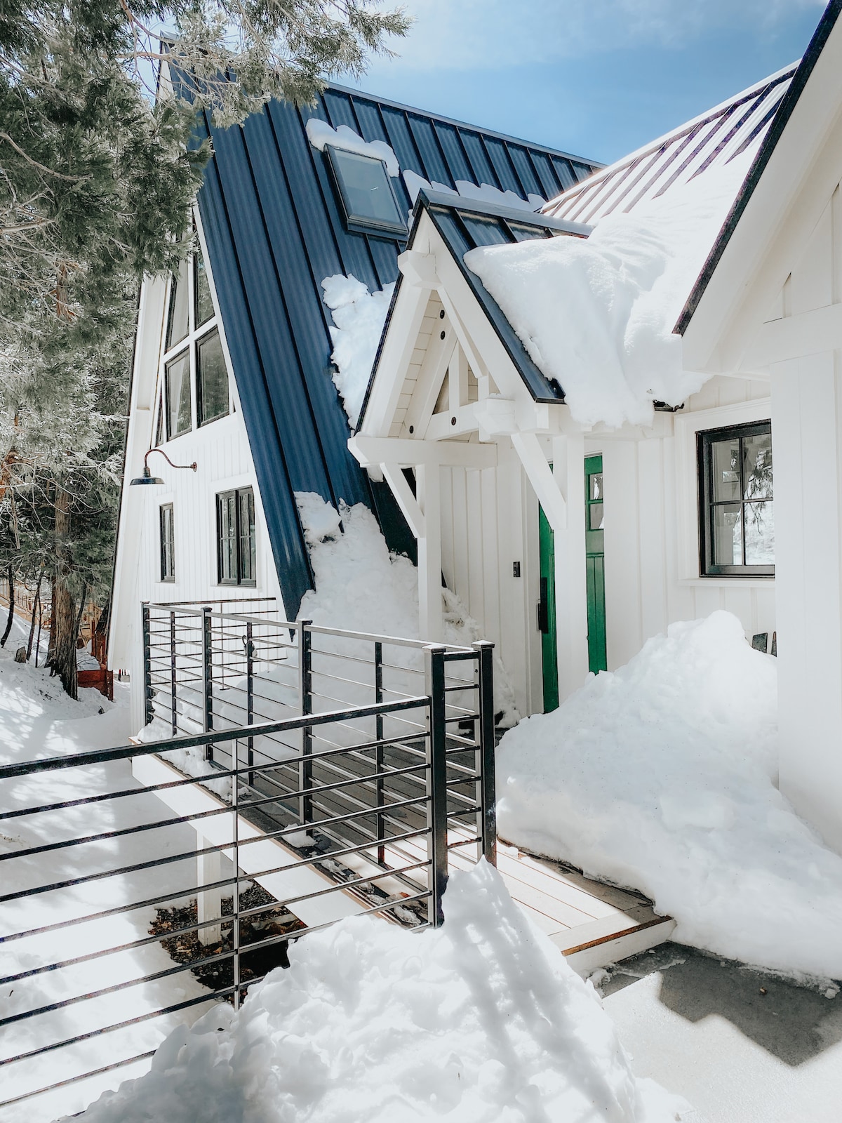 A charming entrance is framed by snow-covered structures featuring a striking combination of navy blue and white siding. A wooden ramp leads to the door, while fresh snow blankets the area, reflecting the sunlight and creating a serene winter ambiance.