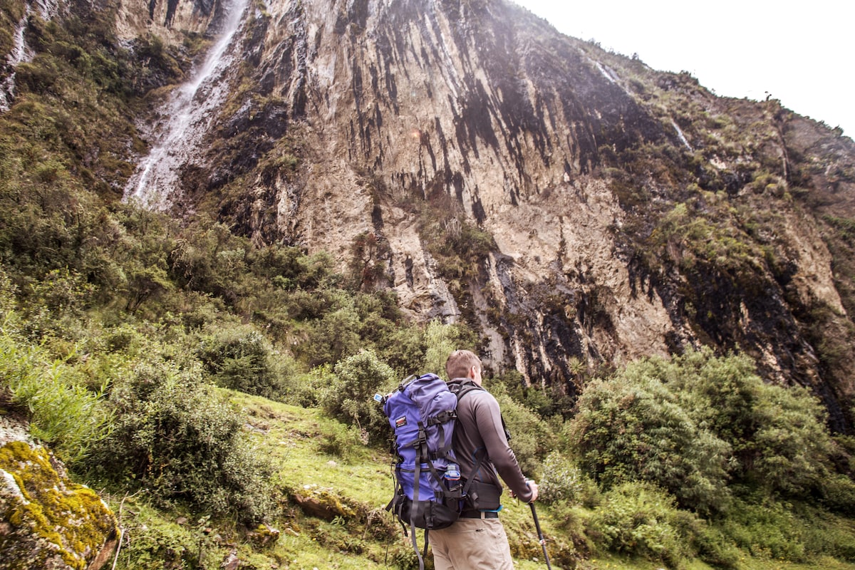 A hiker is seen from behind, standing on a grassy area with lush foliage. A large rocky cliff rises steeply in the background, adorned with patches of vegetation and cascading waterfalls. The scene conveys a sense of adventure in the Andean landscape.