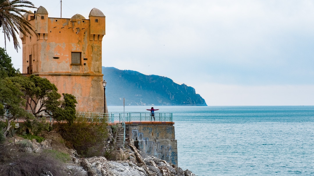 A historic watchtower stands at the edge of the sea, with a person stretching arms towards the horizon. The tranquil water reflects the overcast sky, while the silhouette of hills is visible in the distance. Lush greenery frames the scene, enhancing the coastal landscape.