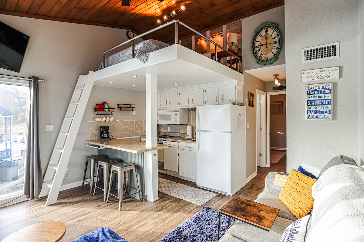 An open living space displays a compact kitchen with white cabinets and modern appliances, complemented by a breakfast bar. A staircase leads to a sleeping loft above. Natural light enters through large glass doors, providing views of the outdoor area.