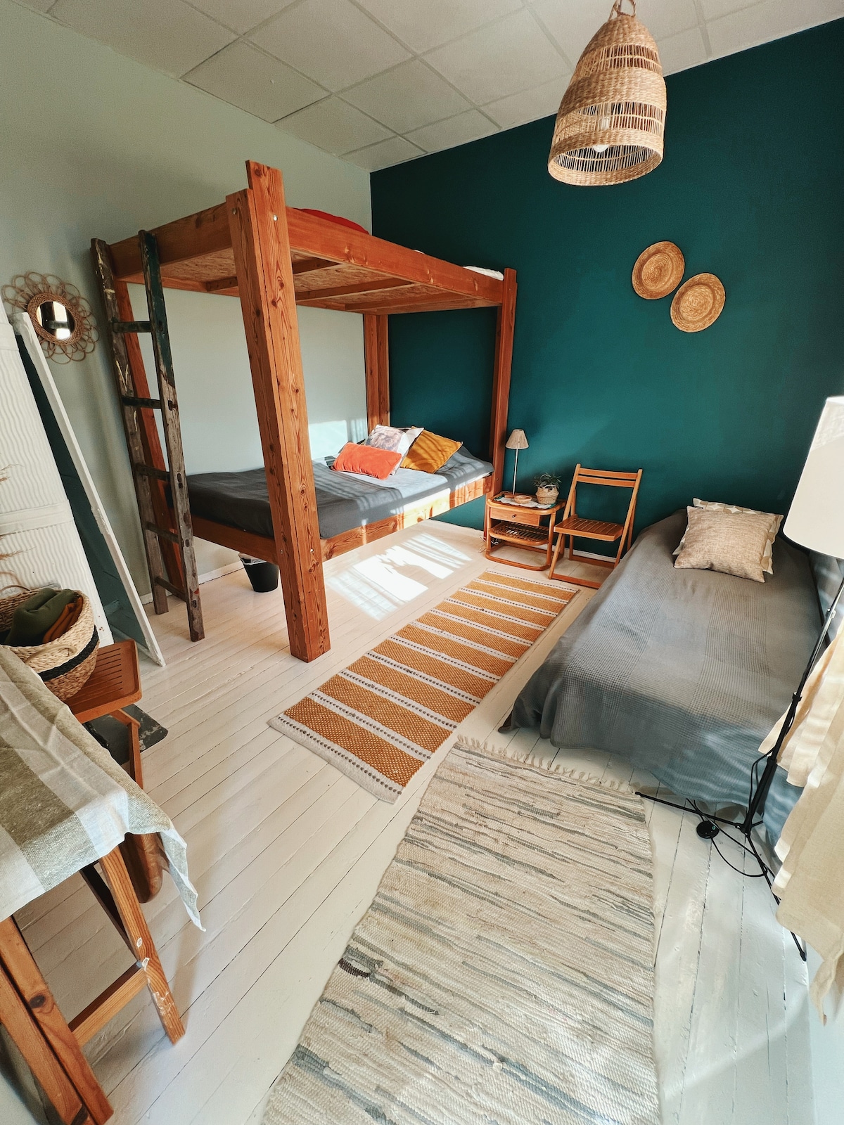The room features a wooden loft bed with a cozy double bed beneath, complemented by a single bed on the opposite side. Textured area rugs and woven wall decor enhance the space, while natural light filters in, creating a bright and inviting atmosphere.