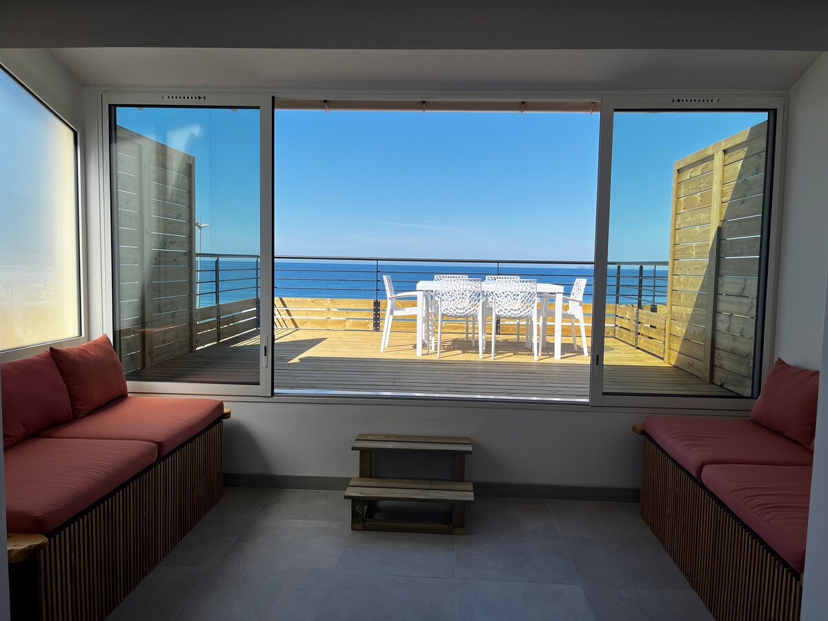 A spacious room features large glass sliding doors that open to a private terrace with outdoor dining furniture. Two built-in seating areas with cushions are positioned near the windows, allowing for a panoramic view of the sea and sky.