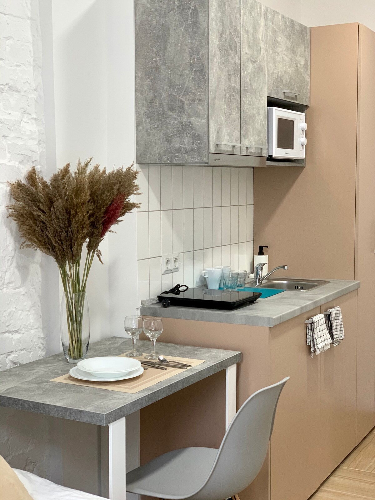 A compact kitchen area showcases modern cabinetry in neutral tones, complemented by a sleek countertop. A small dining table is set with plates and glasses, while a floral arrangement adds a touch of softness. Essential appliances such as a microwave are included for convenience.