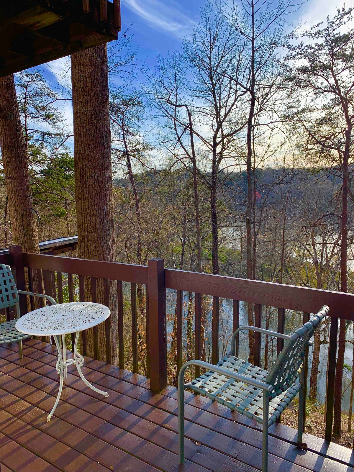 A private deck is shown with a small round table surrounded by two chairs. The railing provides a view of the trees and a river, contributing to a serene natural setting under a clear blue sky.