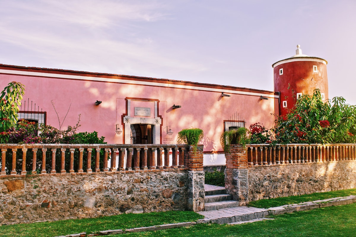 A historic building is adorned with a soft pink facade, paired with a round tower on one side. Lush greenery surrounds the entrance, complemented by a stone wall and well-maintained flower beds. Steps lead to the inviting front door, enhancing the overall charm of the property.