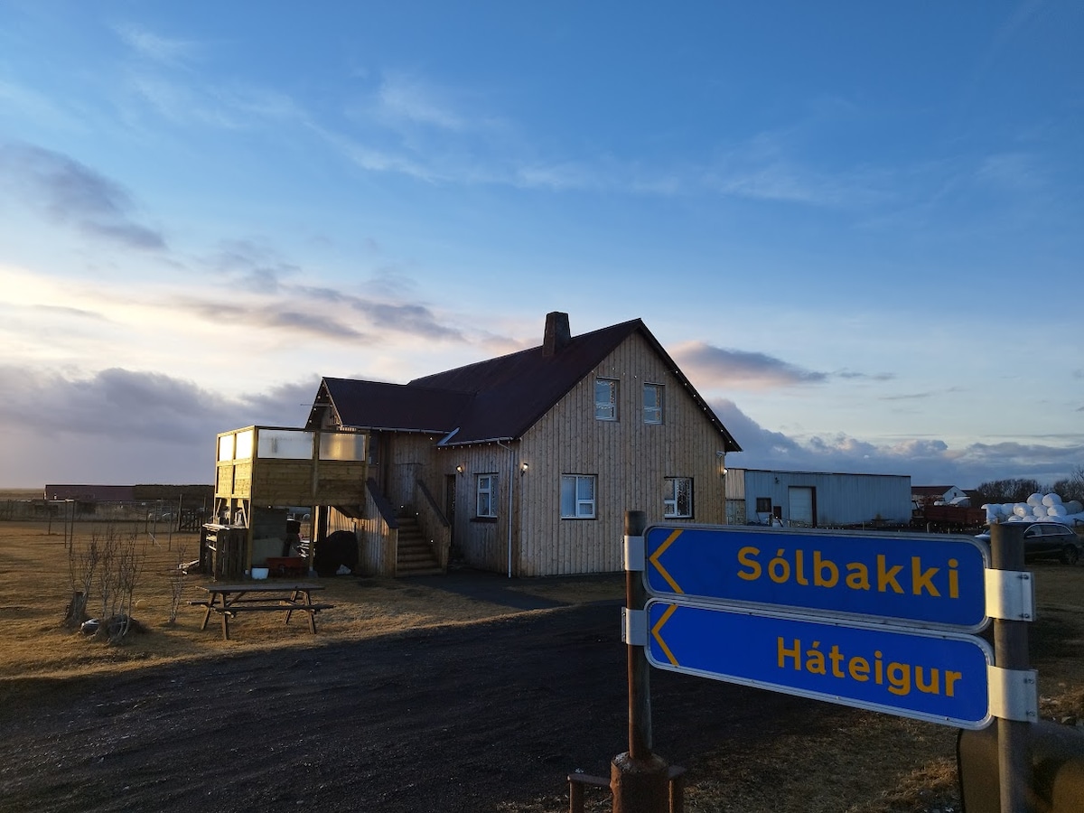 A wooden house with a red roof is positioned against a clear sky, showcasing an entrance with stairs leading to a second-floor guest area. Nearby, a directional sign points towards Sólbakki and Háteigur, with a gravel pathway and open landscapes in the background.