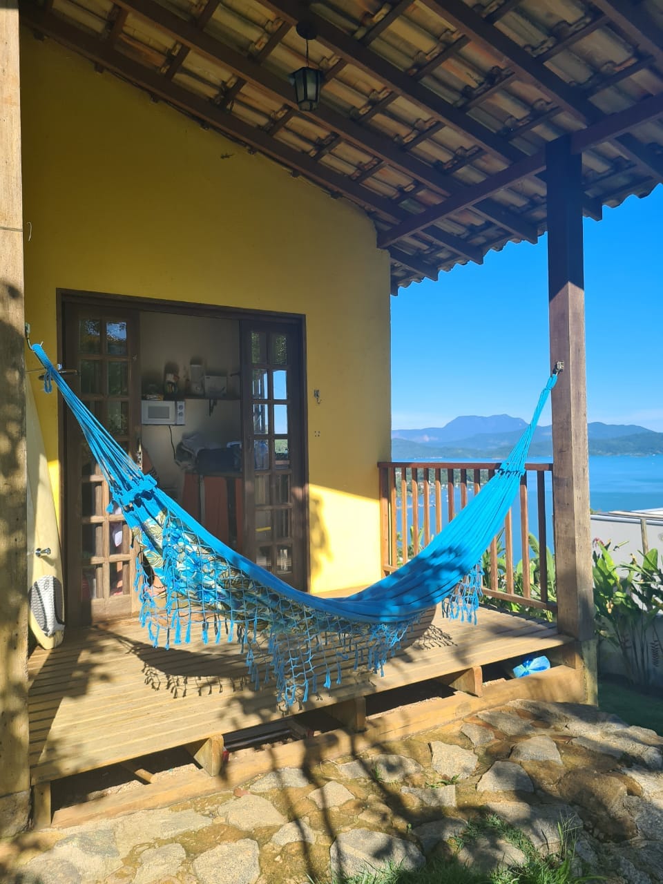 A vibrant blue hammock is suspended from a covered porch, inviting relaxation. Behind it, an open door reveals a cozy interior. The scene features a picturesque view of the sea and distant mountains, framed by rustic wooden beams and a sunlit sky.