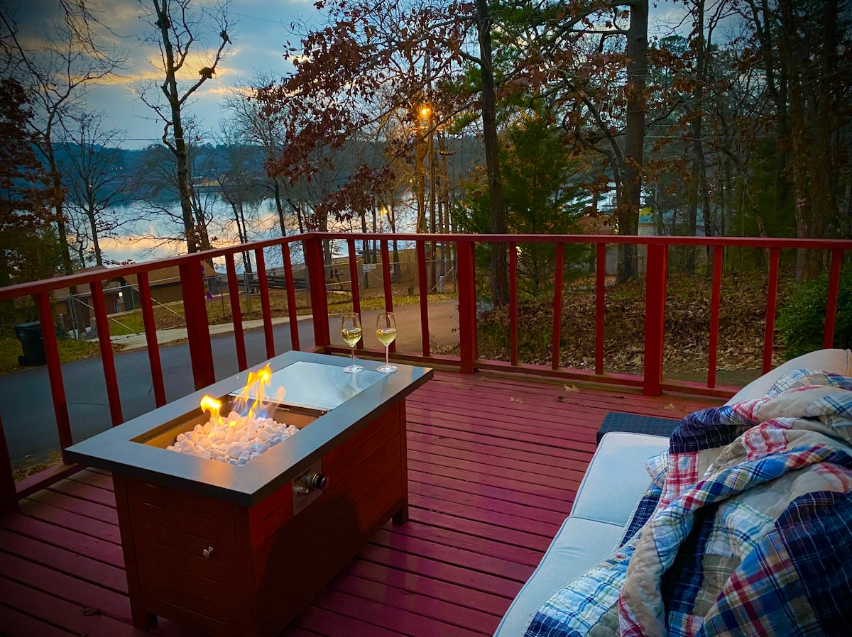 A spacious deck features a modern fire pit table with flames dancing over ice, accompanied by two glasses positioned nearby. A cozy throw blanket lies draped over a relaxed seating area, complemented by a serene view of the lake and surrounding trees at dusk.