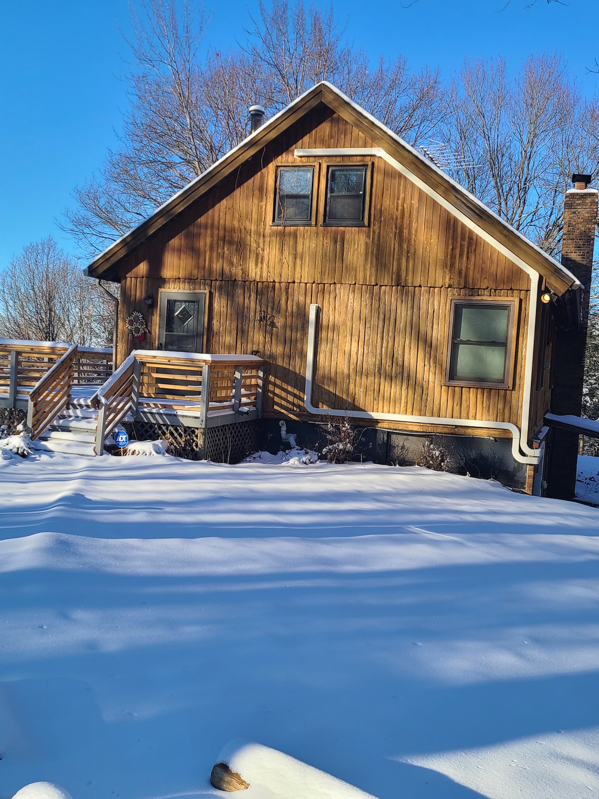 A wooden cabin is framed by snow-covered ground, highlighting the natural beauty of its surroundings. The structure features multiple windows and a set of stairs leading to the entrance. Tall trees are visible in the background against a clear blue sky.