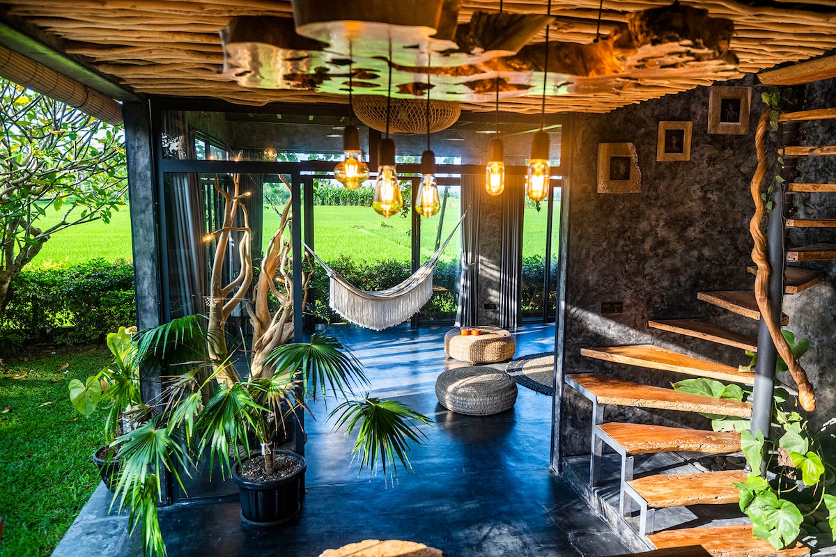 The common area features an inviting open layout with a blend of natural elements and modern design. Hanging pendant lights illuminate the space, while a hammock and various plants add a touch of greenery. Large glass doors provide views of the surrounding rice fields.
