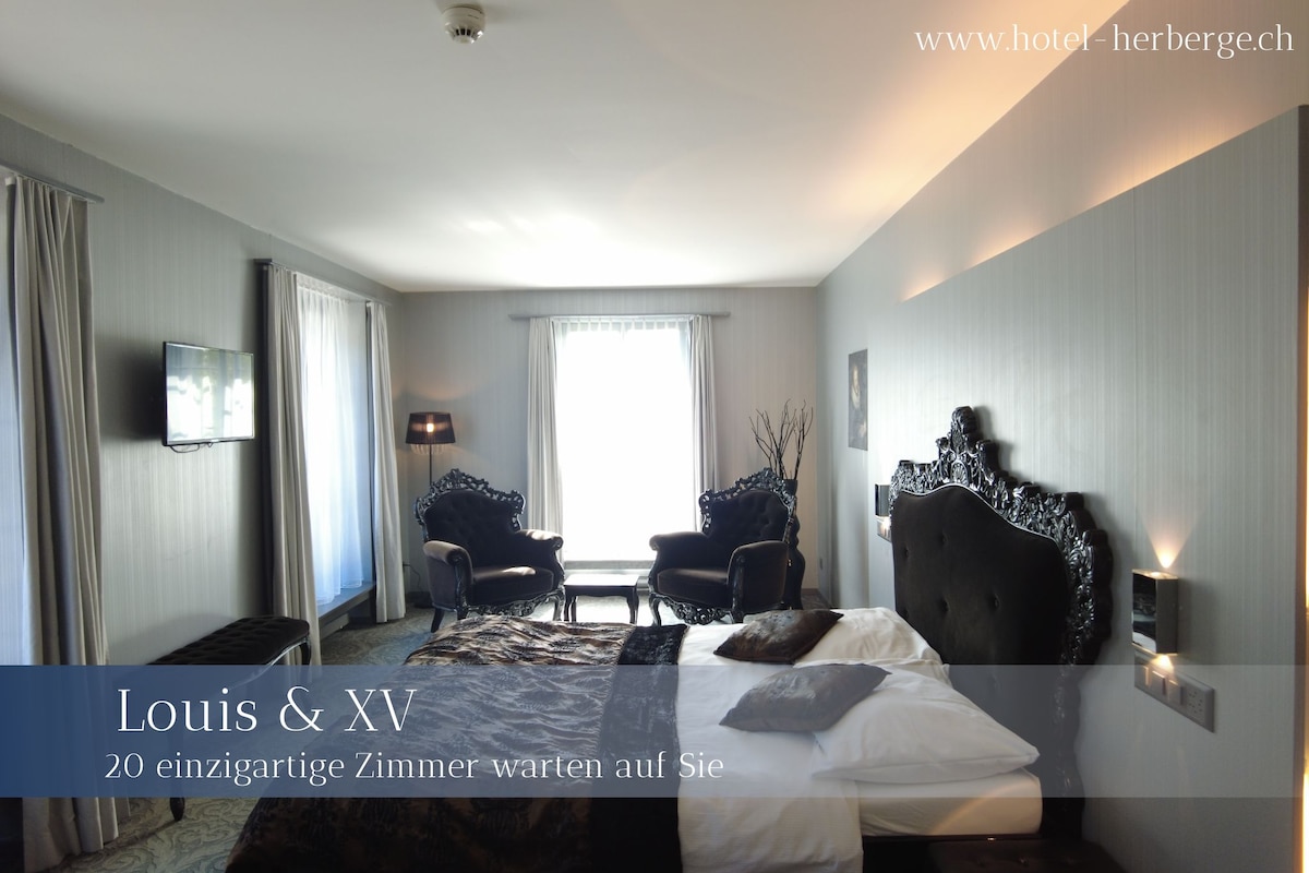 A modern room features stylish dark furnishings and intricate wall decor. A spacious bed is centered in the area, accompanied by two elegant chairs. Natural light is welcomed through large windows, enhancing the serene ambiance of the space.