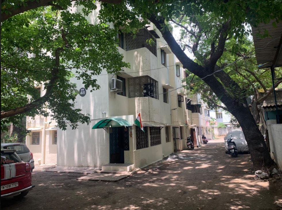 The view showcases a multi-story building surrounded by lush trees, providing ample shade. A neatly paved area with designated parking spaces is visible directly in front. The entrance features a green awning, along with nearby bicycles and a welcoming atmosphere.