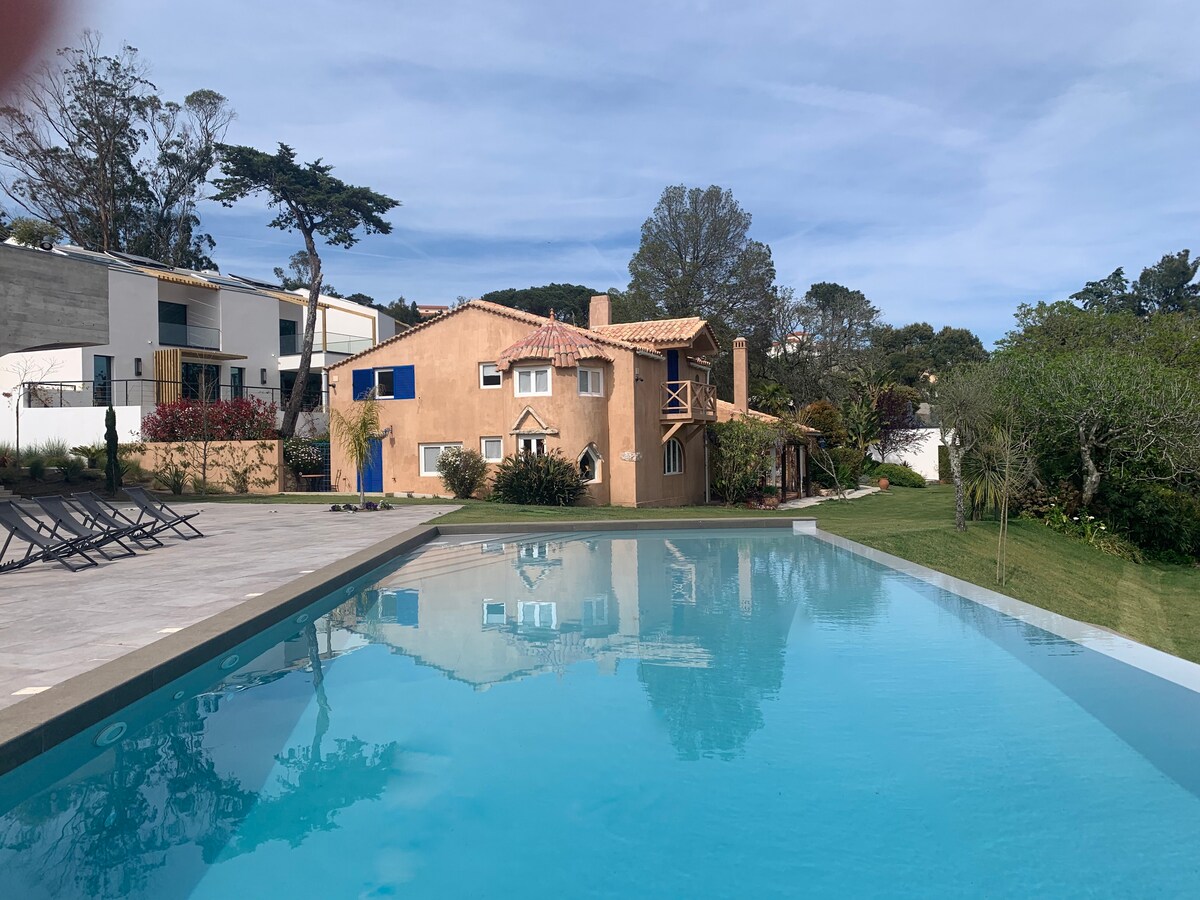 The expansive infinity pool reflects the serene blue sky, bordered by a sunlit terrace and loungers. In the background, a two-story villa with a warm terracotta facade is surrounded by lush greenery and trees, creating a peaceful outdoor setting.