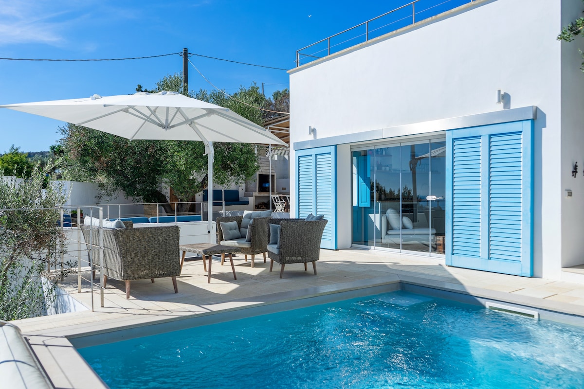 A private pool surrounded by a spacious terrace is highlighted, featuring a patio with wicker chairs and a large umbrella for shade. Large glass doors provide access to an indoor living space, framed by light blue shutters that complement the overall light décor.