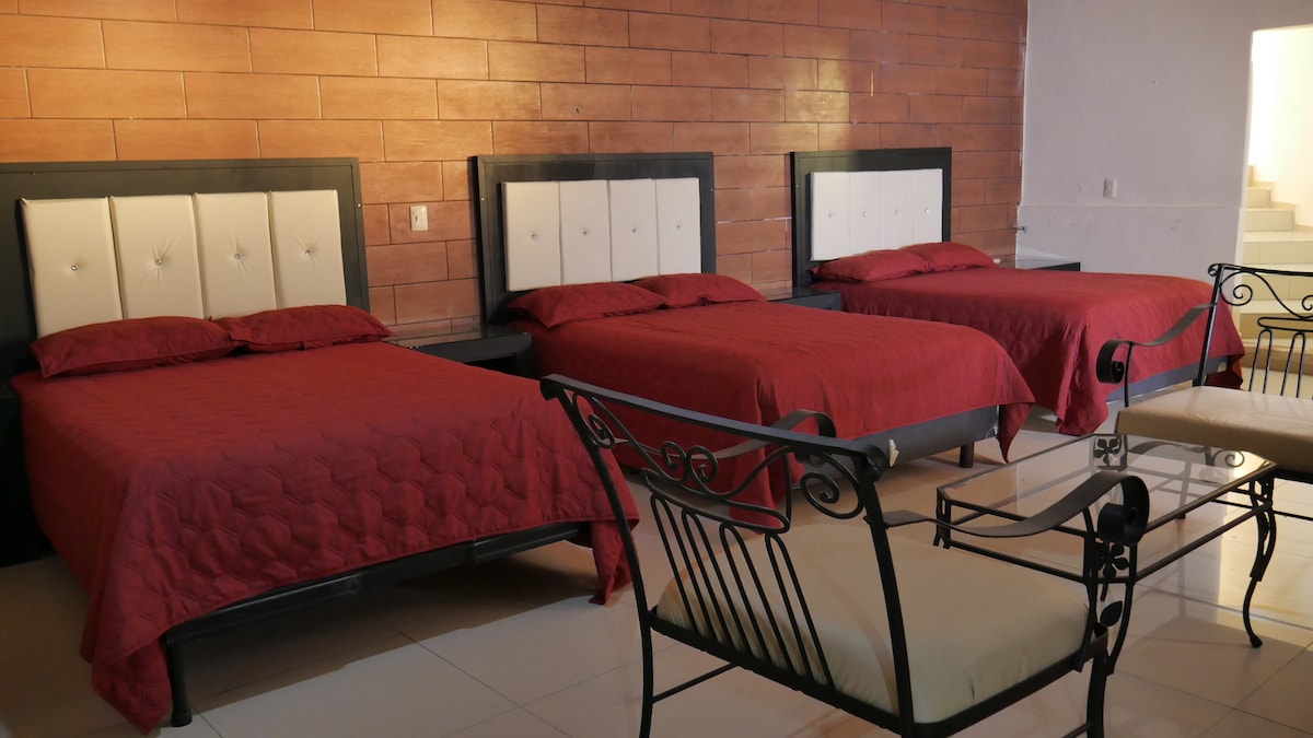 A spacious sleeping area features three beds with burgundy covers and padded headboards, offering ample space for guests. A seating arrangement with a glass-top table and wrought iron chairs is positioned nearby, providing an inviting spot for relaxation.