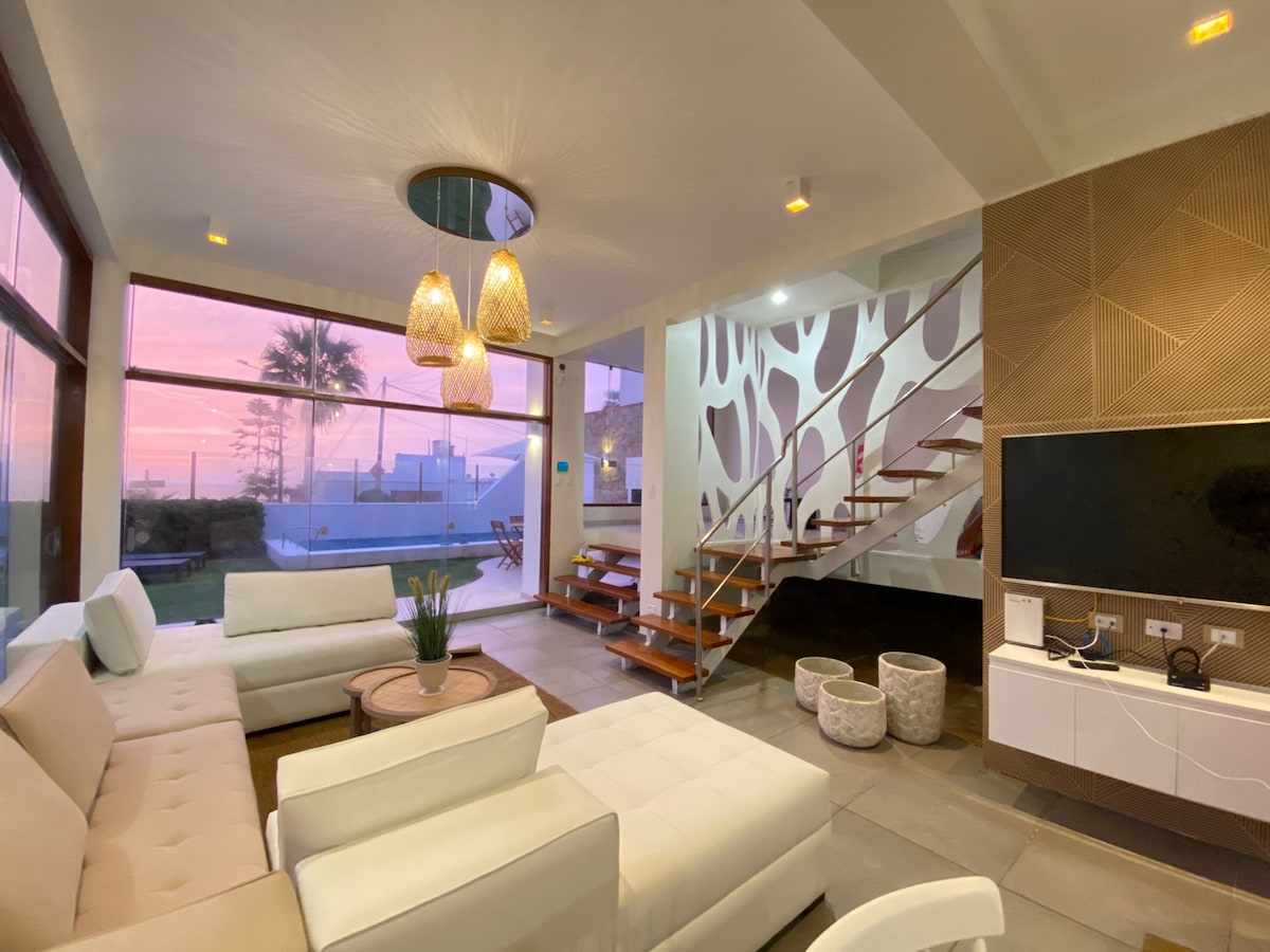 An open and airy living space features large windows that provide a view of the exterior landscape. Stylish white furniture is arranged for comfort. A modern staircase leads to the upper level, and soft lighting creates a welcoming ambiance.