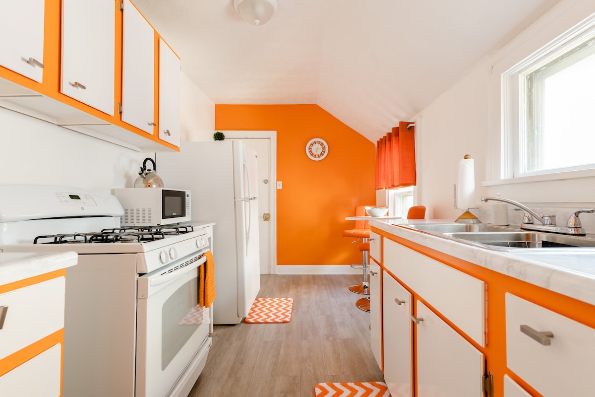 A bright kitchen features vibrant orange accents on the walls and cabinetry. Modern appliances include a gas stove, microwave, and refrigerator. The space is complemented by cheerful window treatments and coordinating floor mats, creating a lively atmosphere for cooking and dining.