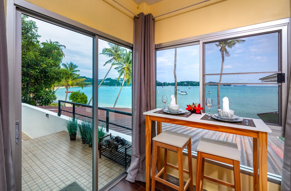A coastal dining area features a high table with two stools, positioned next to large glass windows. A view of the sea and palm trees is visible, highlighting the tranquil waters with several sailboats in the distance.