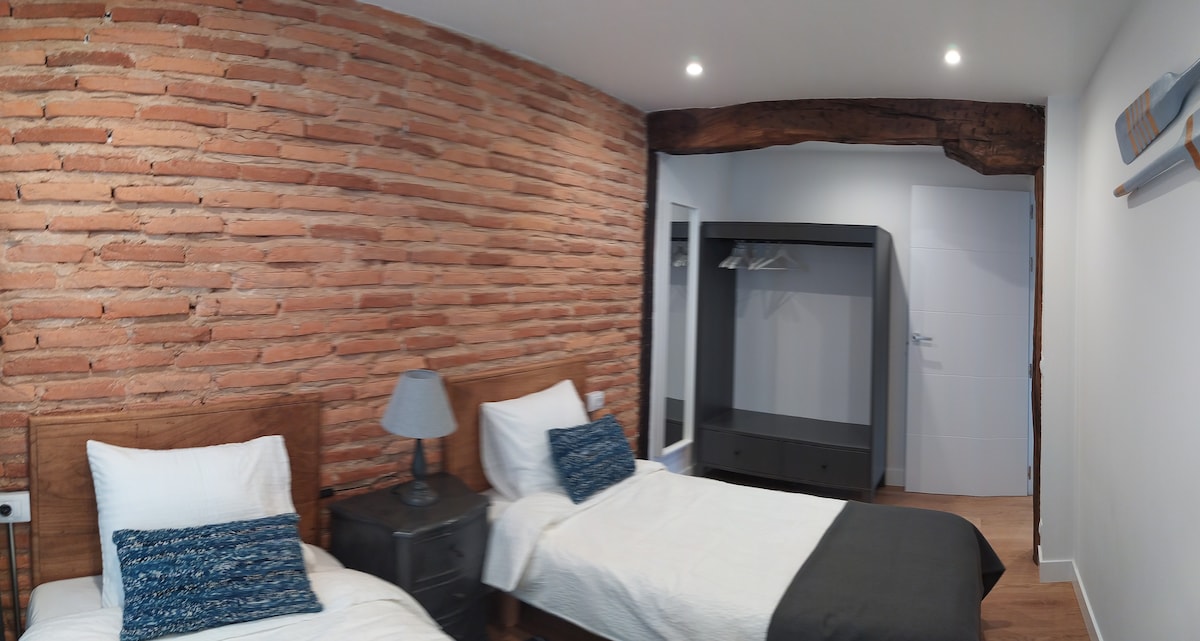 חדר שינה עם 2
מיטות זוגיות וקיר לבנים חשוף לאופי