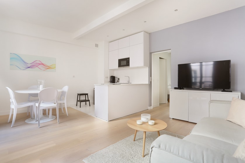 A bright living area displays light wooden flooring with a plush area rug. A modern kitchen with white cabinetry is visible, alongside a dining table with four white chairs. A comfortable sofa is positioned near a large television, offering a cozy space for relaxation.