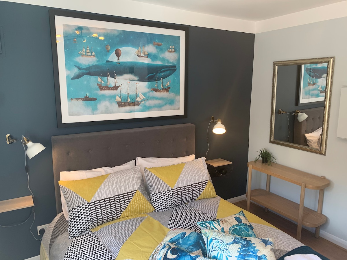 A cozy bedroom is highlighted by a framed artwork featuring a blue whale and various ships. The bed is adorned with a patchwork of yellow and gray patterned bedding. Two bedside lamps are mounted on the wall, and a mirror reflects the room's inviting ambiance.
