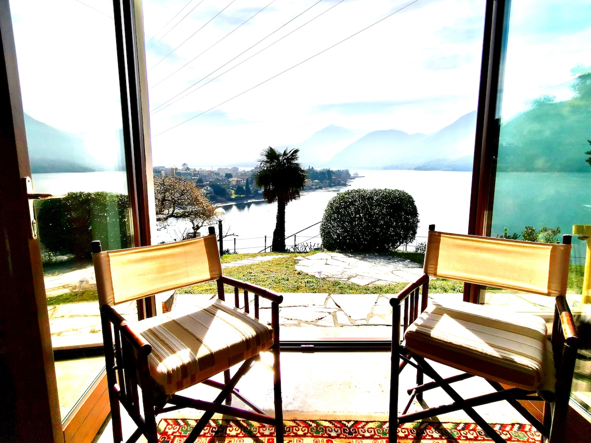Two wooden chairs are positioned facing a large glass door, providing an unobstructed view of the lake and surrounding mountains. Soft light filters through, highlighting the serene landscape outside, while a patterned rug adds a touch of warmth to the interior space.
