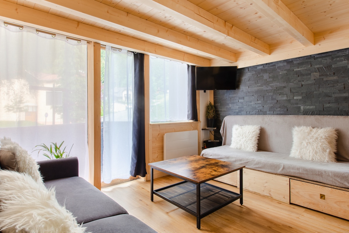 A bright living area features large windows draped with sheer curtains, allowing natural light to fill the space. A modern coffee table stands centered on the wood floor, complemented by a plush sofa with decorative pillows. A flat-screen TV is mounted on a stylish stone wall.