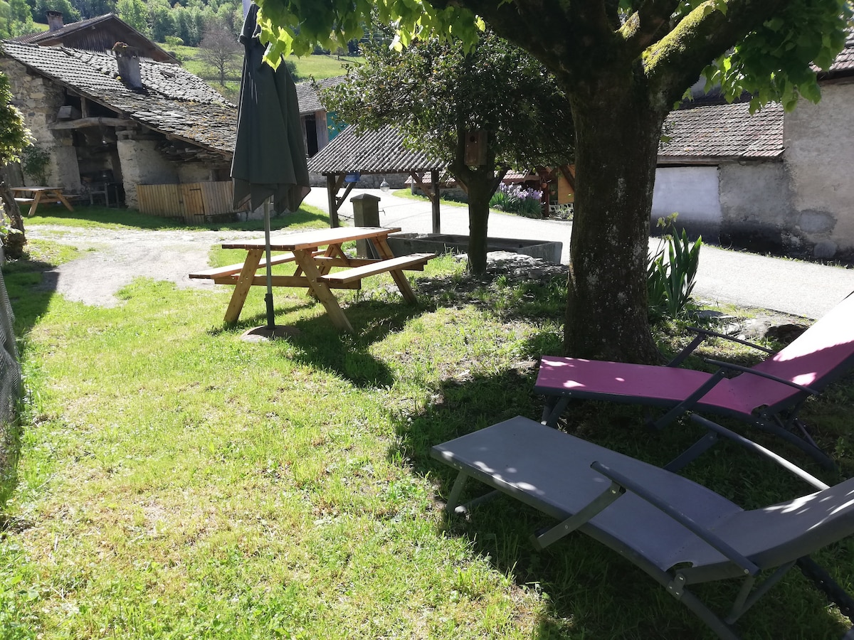 A grassy outdoor space is shown, featuring two reclining lounge chairs in shades of pink and grey. A picnic table is positioned nearby under the shade of a tree, with a large umbrella providing additional shelter. The scenic area is bordered by rustic stone structures.
