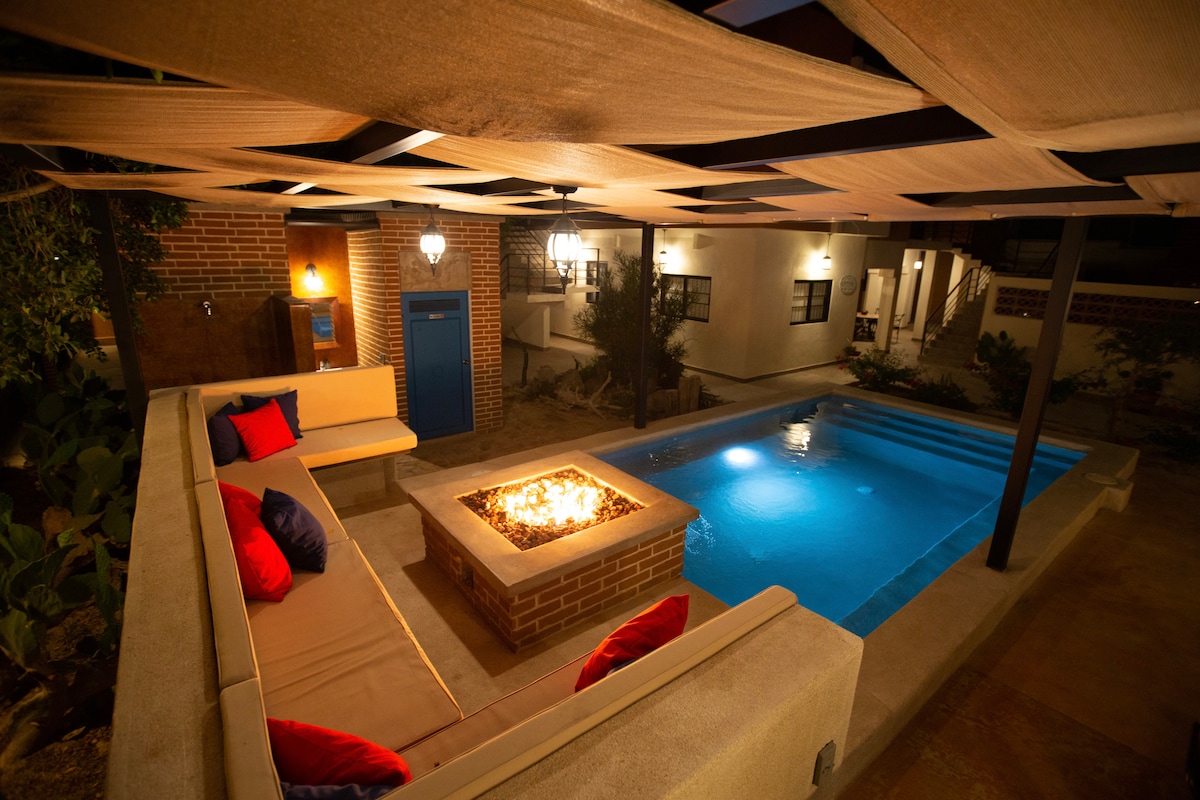 A well-lit outdoor area features a sparkling swimming pool and a seating arrangement surrounding a fire pit. Soft seating with cushions in vibrant colors offers comfort under a shaded canopy. Lush greenery and light bricks create a calming atmosphere in the shared space.