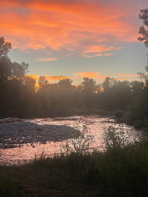 A serene river reflects the warm hues of a sunset, framed by lush greenery and trees along the banks. The gentle flow of water is visible, accompanied by smooth stones that create a natural landscape, inviting peaceful moments outdoors.