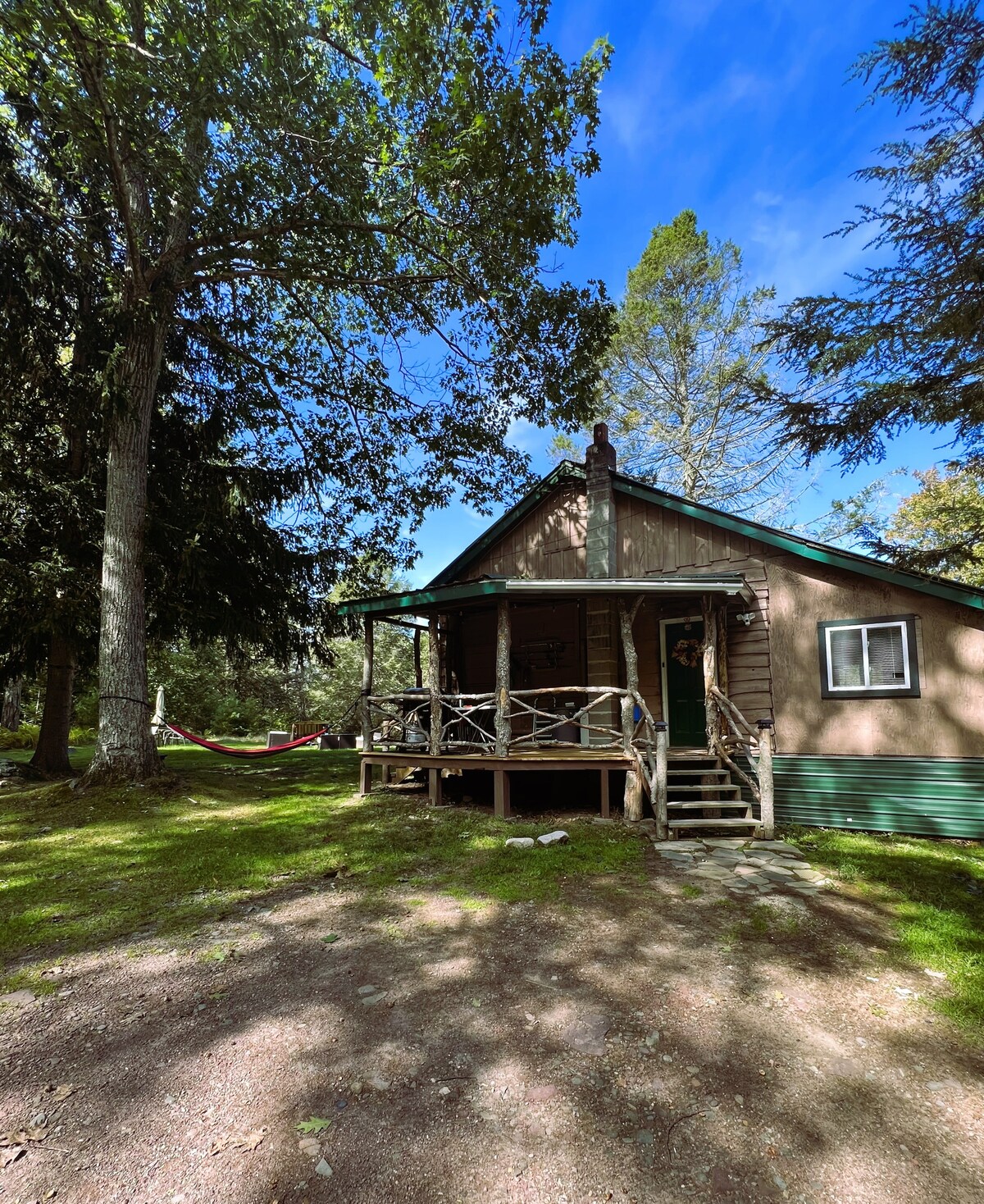 A rustic cabin is set amidst a lush green yard, with trees providing shade. The entrance features a small porch with steps leading up to the door, complemented by a hammock strung between two trees nearby.