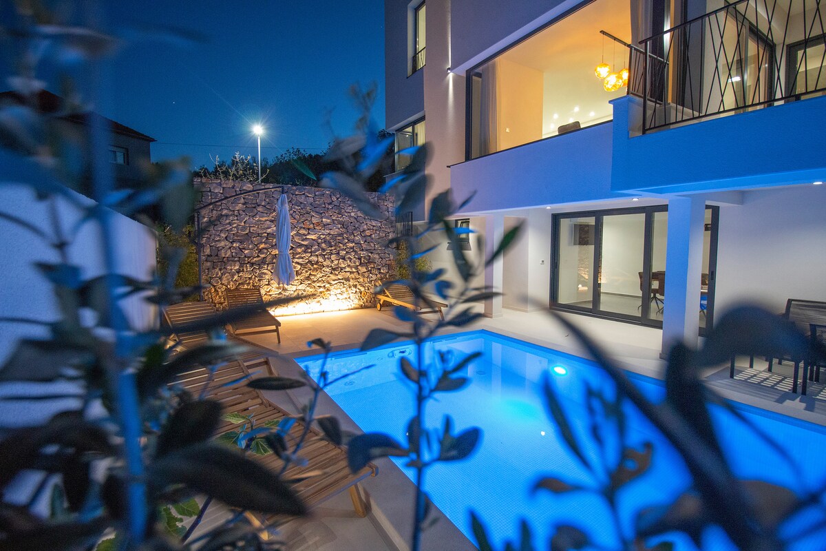 An outdoor area is illuminated softly at night, featuring a clear swimming pool surrounded by wooden lounge chairs. The light showcases a charming stone wall, while large glass doors lead into the villa's interior, highlighting the seamless connection between indoor and outdoor spaces.