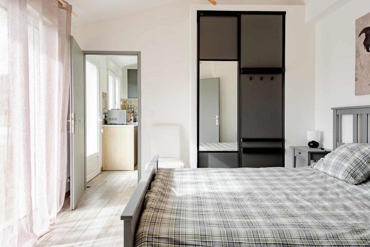 A well-lit bedroom features a spacious bed covered with a plaid blanket. A sliding glass door leads to the kitchen area, with additional natural light filtering through sheer curtains. A wardrobe is present, and subtle décor complements the minimalist design.