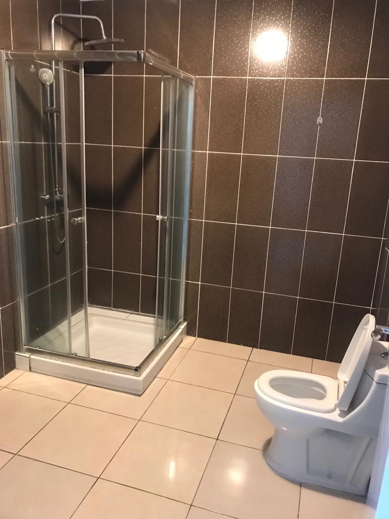 An enclosed bathroom space is shown, featuring a glass shower with chrome fixtures on one side. A modern toilet is positioned adjacent to the shower. The walls are adorned with dark tiles, and light-colored tiles cover the floor.
