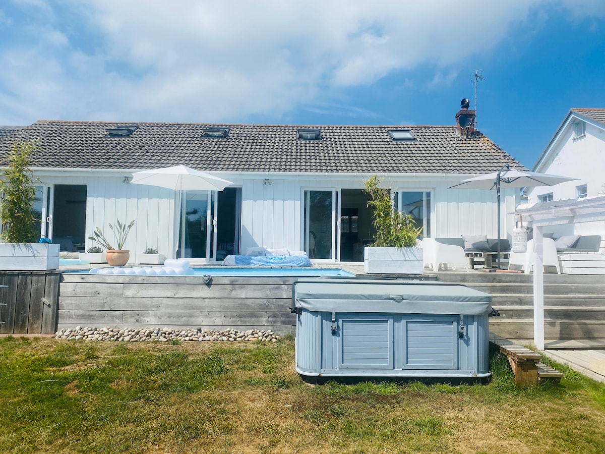 The exterior of a modern home features a hot tub with a stylish wooden surround. Sun loungers and parasols are arranged neatly by a spacious terrace, while large glass doors reveal a bright and airy indoor living area. Green grass adds to the inviting appeal.