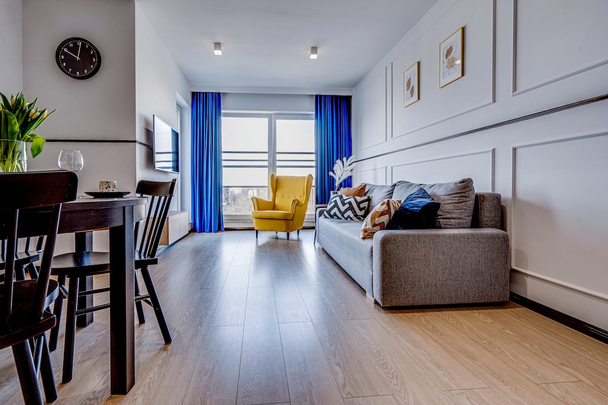 A modern living area features a grey sofa with decorative pillows, a yellow armchair, and a dining table with chairs. Natural light enters through large floor-to-ceiling windows, enhanced by blue curtains. A clock hangs on the wall above the television, adding a functional touch.