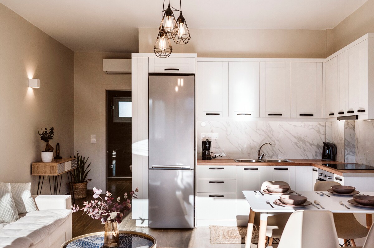 A modern kitchen area features white cabinets paired with wooden countertops. A stainless steel refrigerator stands prominently, and a dining table is set with neutral dishware. Minimalist decor includes a small vase with flowers and ambient lighting from stylish pendant fixtures.