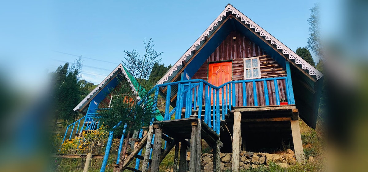Two cozy cottages are positioned on a gentle slope, surrounded by lush greenery. The structures feature sloped roofs and vibrant blue accents on the balconies and staircases, inviting guests to enjoy the serene natural surroundings.