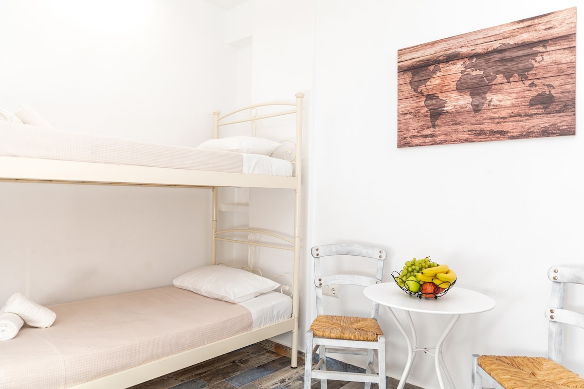A light-infused room features a bunk bed with a simple design. A single bed sits below, while a round dining table is accompanied by two chairs. A wooden wall art depicting a world map adds a global touch to the decor.