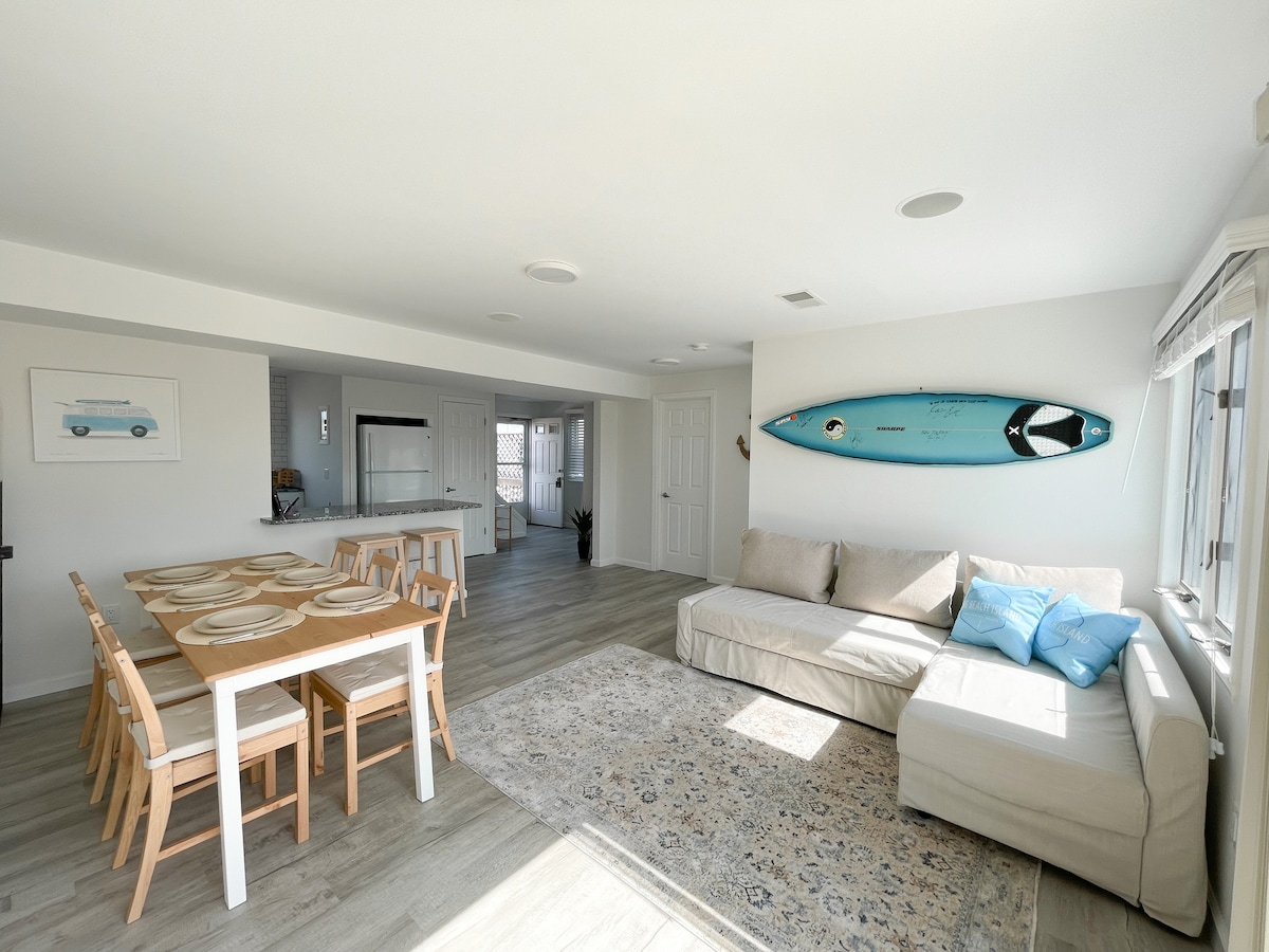 An open-concept living space features a light-colored sofa adorned with blue accents and a dining table set for eight. A surfboard is mounted on the wall above, while large windows allow natural light to fill the area, enhancing the airy feel of the room.