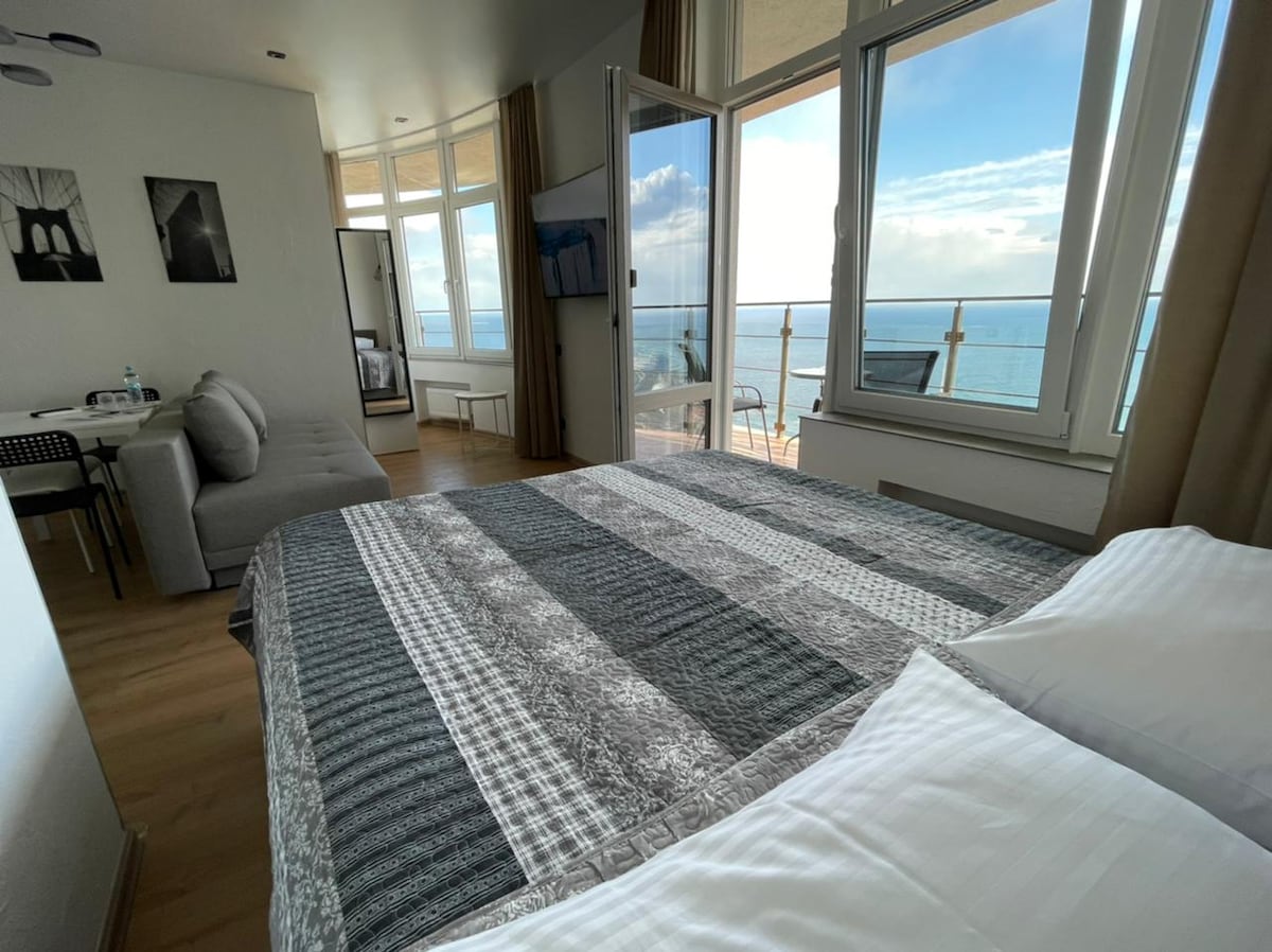 A comfortable sleeping area features a spacious bed with a patterned blanket. Large windows frame a view of the sea, while a sofa and dining table create an inviting living space. Natural light brightens the room, enhancing the overall ambiance.